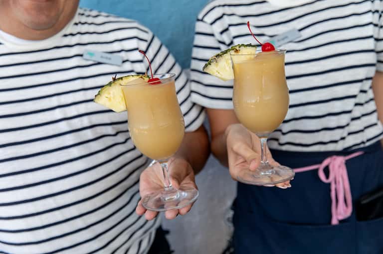 Two tropical cocktails with pineapple garnish held by people wearing navy and white striped shirts