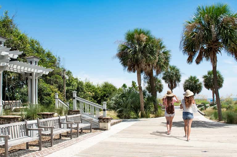 Wooden boardwalk with palm trees and beachgoers in wide-brimmed hats