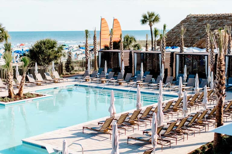 Beachfront resort pool with palm trees, white umbrellas, lounge chairs, and ocean view