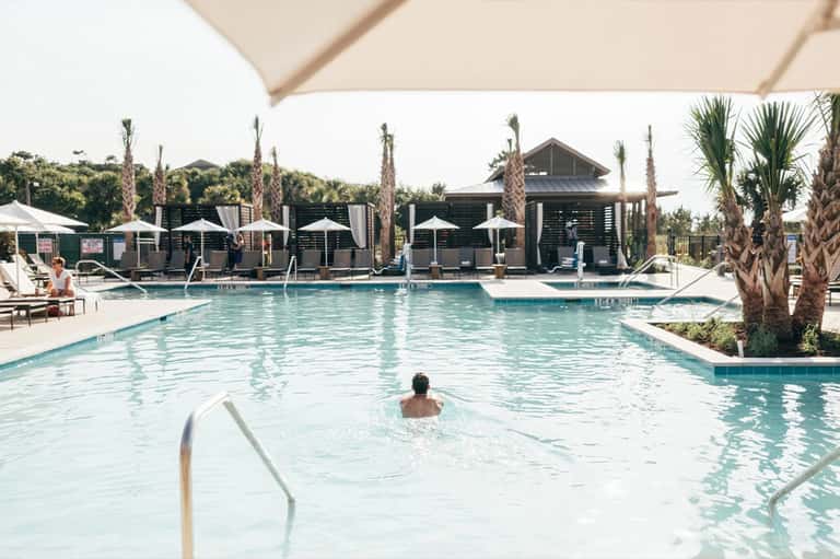 Daytime resort pool with guests swimming, palm trees, white umbrellas, and poolside cabanas