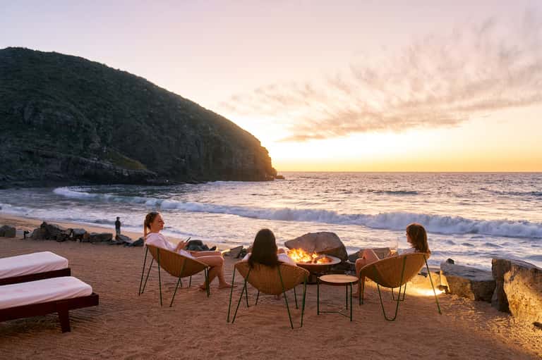 Four people sitting around a beach bonfire at sunset with ocean and rocky cliffs