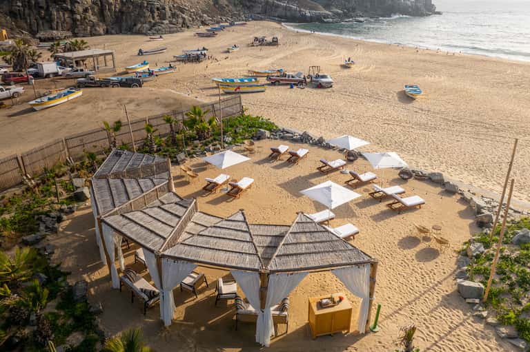 Beach scene with wooden cabanas, white umbrellas, boats, and rocky cliffs in background
