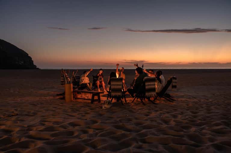 Group of people gathered around bonfire on beach at sunset with chairs and desert backdrop