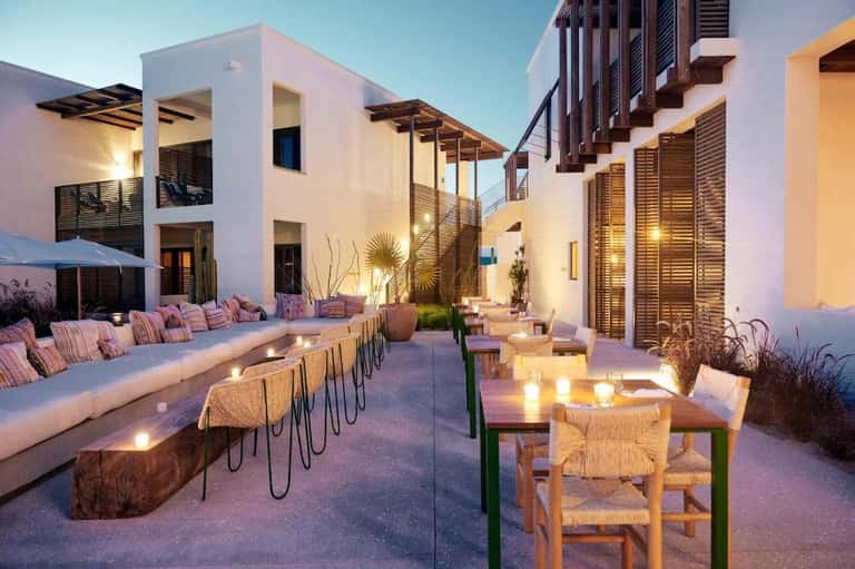 Contemporary resort courtyard at dusk with white buildings, pink striped cushions, lounge seating, and warm lighting
