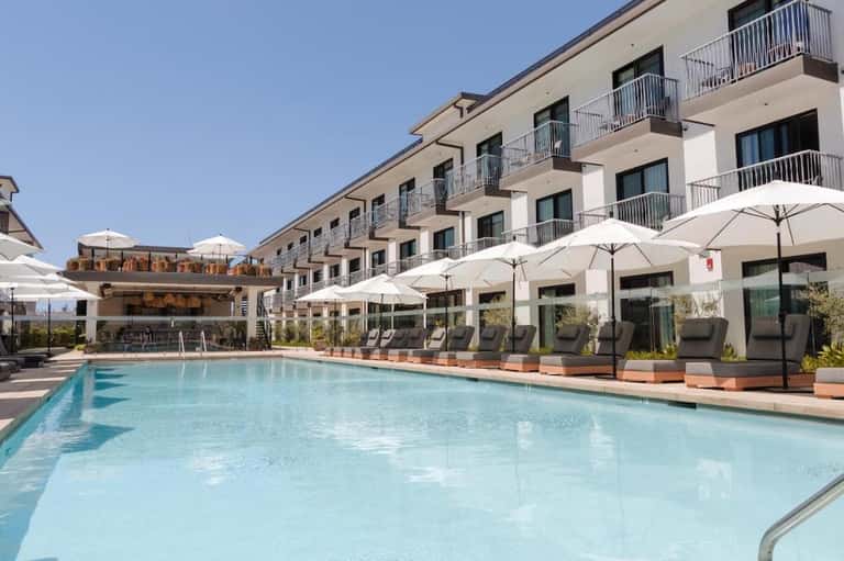Outdoor pool with light blue water, white umbrellas, lounge chairs, and multi-story hotel building