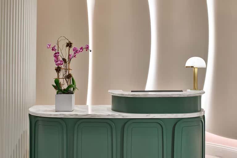 Elegant reception desk with curved green cabinetry, marble top, white lamp, and orchid flower arrangement