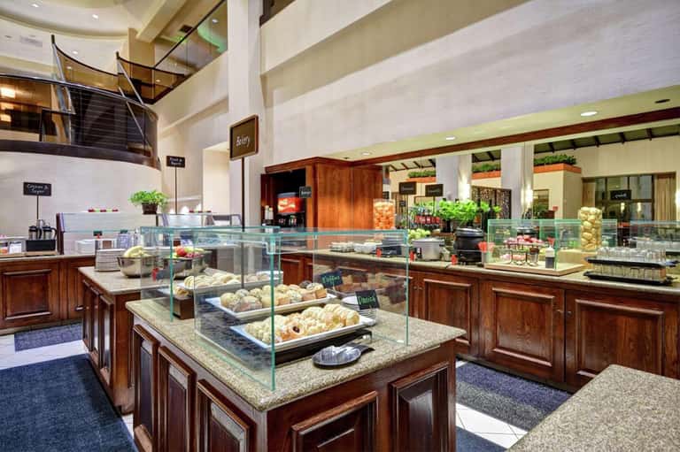 Upscale hotel bakery with glass display cases showcasing pastries and baked goods, wood cabinetry, and multi-level seating area