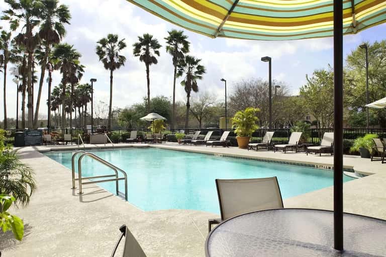 Resort pool area under large shade structure with palm trees, lounge chairs, and umbrella in tropical setting