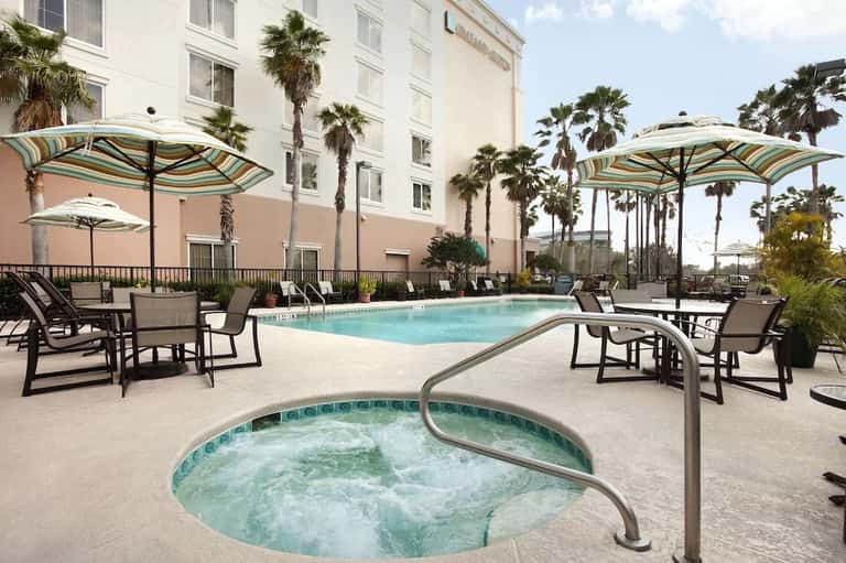 Resort pool deck with striped umbrellas, lounge chairs, hot tub, and palm trees with waterfront views