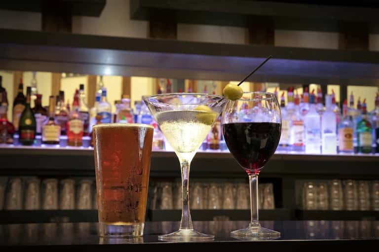 Three cocktails on bar counter: cola drink, martini with olive, and red wine with bottles behind