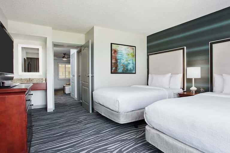 Double queen hotel room with striped bedding, red accent desk, and bathroom entrance