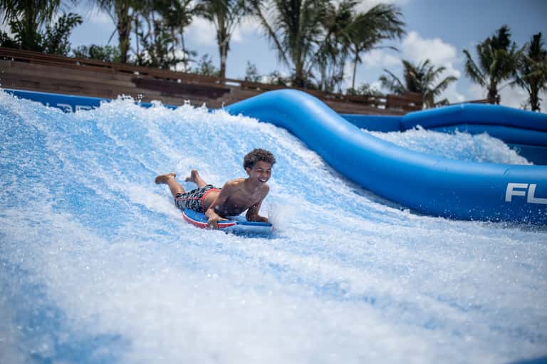 Child body-surfing on artificial wave machine with blue slide and palm trees in background