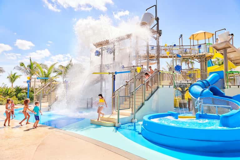 Waterpark with splash pad structure, water bucket, slides, children playing, and palm trees under blue sky