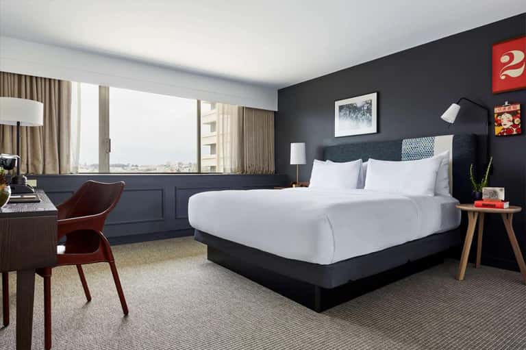 Contemporary hotel bedroom with dark accent wall, white bedding, floor-to-ceiling windows, and city skyline view