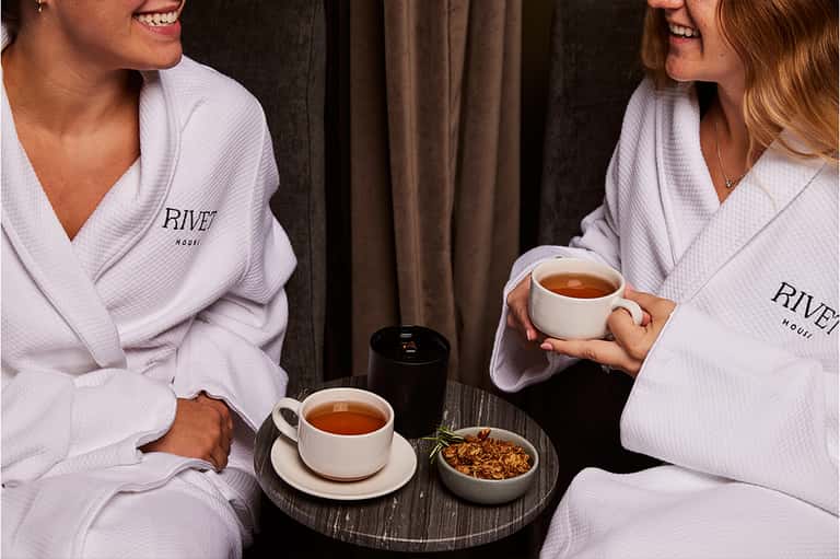 Two women in white robes enjoying tea and snacks at a spa relaxation area