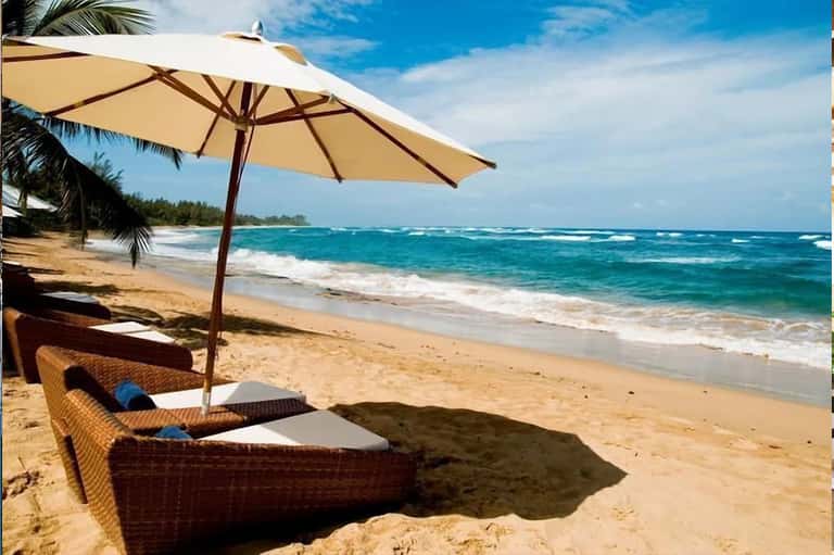 Beach lounge area with tan umbrella, rattan chair, and turquoise ocean waves in background