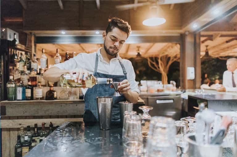 Bartender in denim apron pouring cocktail at modern bar with bottles and glassware.