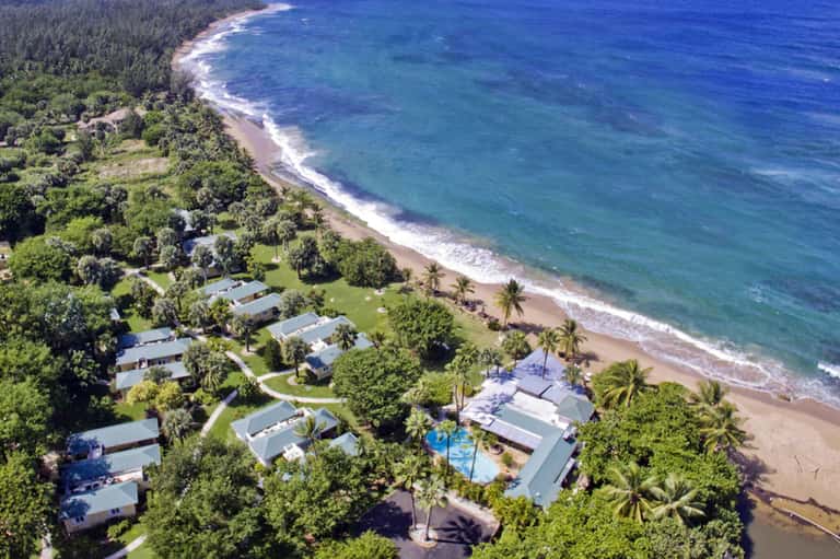 Aerial view of beachfront resort with pool, cabins, palm trees, and pristine beach and ocean