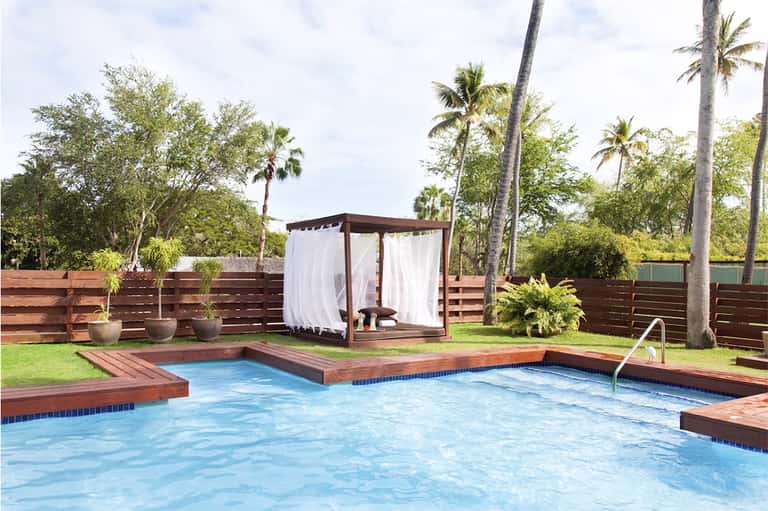 Residential pool with wood decking, brown cabana with white curtains, palm trees, and fenced tropical setting