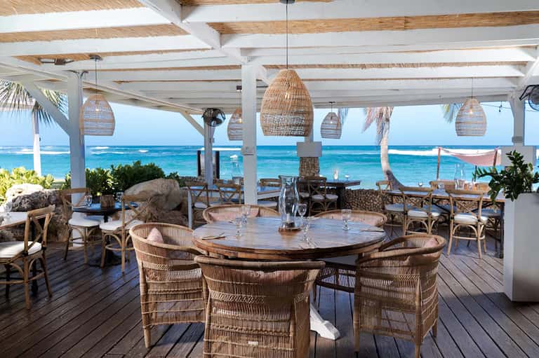 Beachfront dining area with woven pendant lights, wooden tables, ocean view, and palm trees