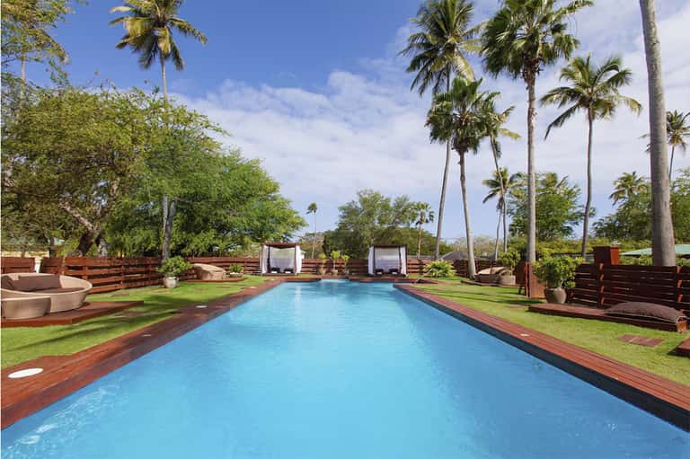 Resort pool surrounded by palm trees, green lawn, wooden deck, and cabana pavilions