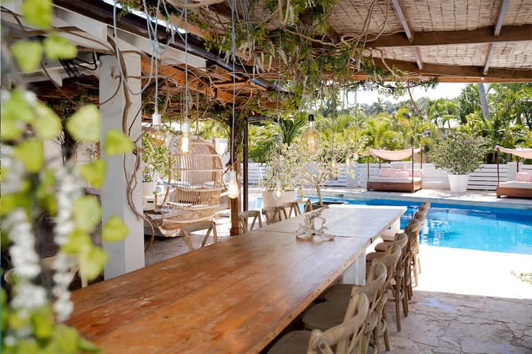 Covered poolside dining area with wooden table, hanging plants, and blue pool view