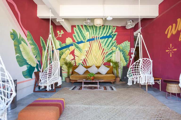 Tropical lounge area with hanging white chairs, banana leaf mural, wooden sofa, and colorful decor