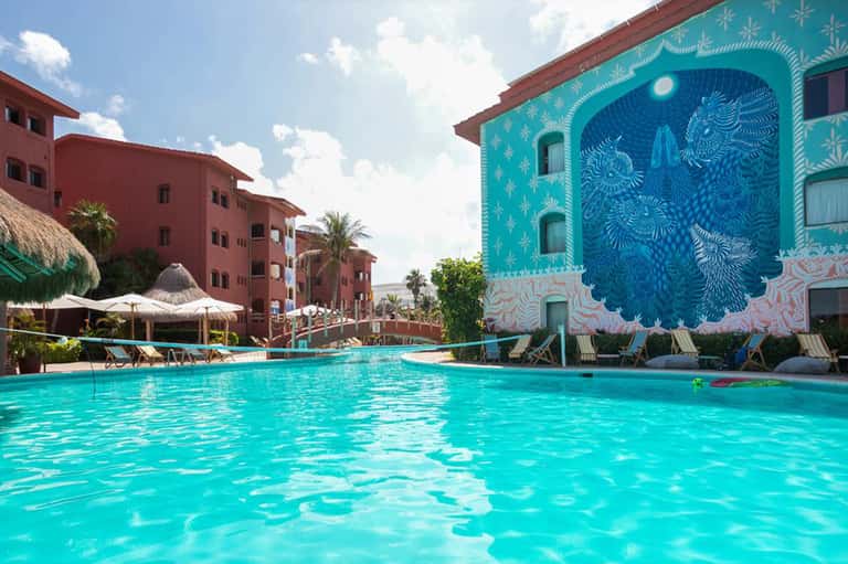 Turquoise resort pool with colorful mural wall, lounge chairs, and white umbrellas