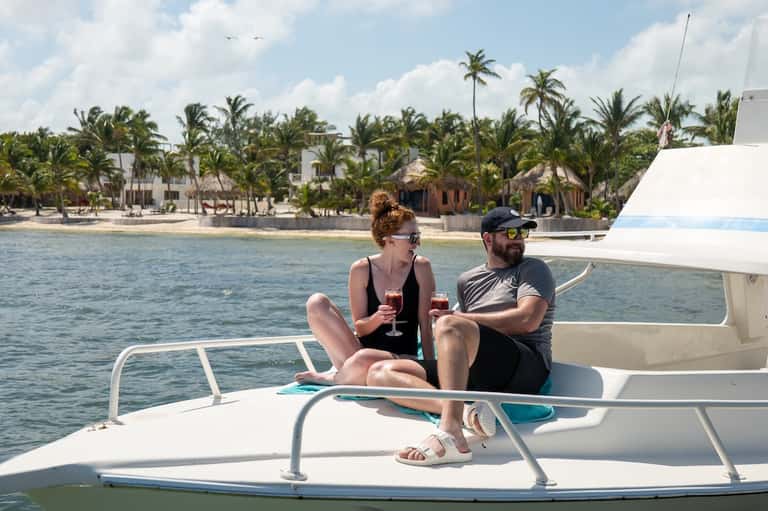 Couple enjoying drinks on a boat with palm-lined beach resort visible in background