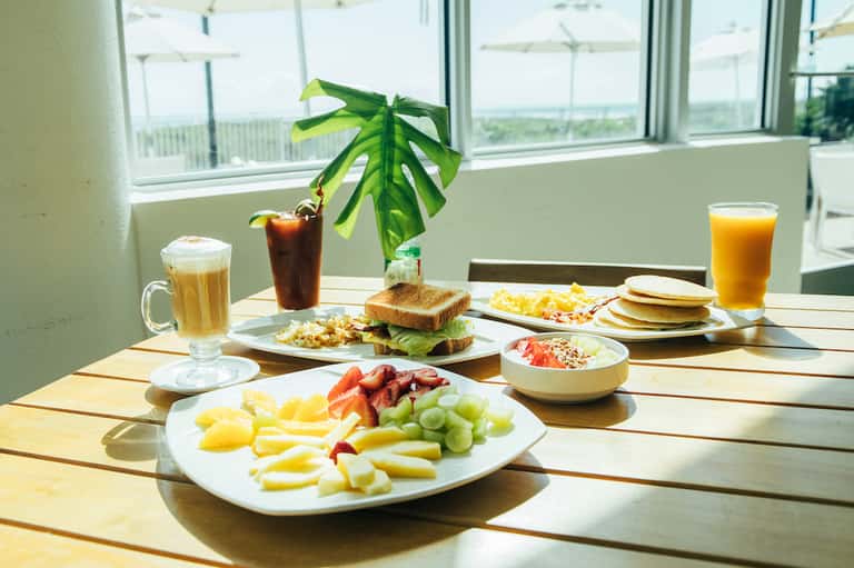 Breakfast spread with fresh fruits, bacon, pancakes, beverages, and potted plant on sunny dining table