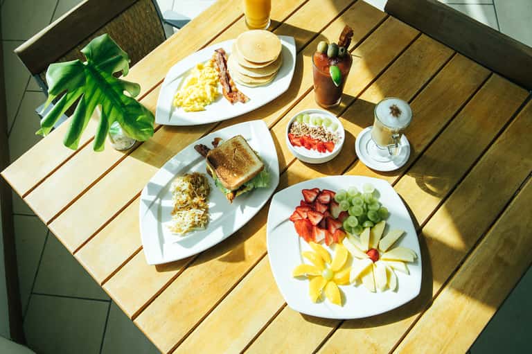 Breakfast spread on wooden table with pancakes, eggs, bacon, fresh fruit, and beverages