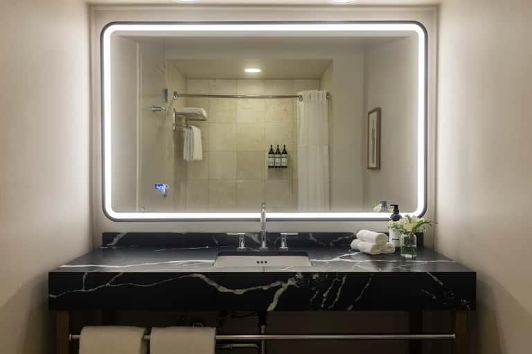 Luxurious bathroom with large illuminated mirror, black marble vanity, glass shower enclosure, and premium toiletries