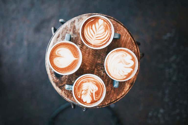 Four latte cups with decorative latte art arranged on a round wooden table, overhead view