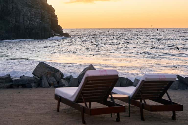Two lounge chairs on sandy beach at sunset with rocky cliffs and ocean backdrop