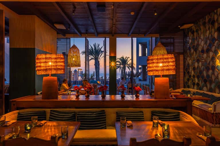 Upscale restaurant with woven pendant lights, palm tree views, and sunset ocean backdrop through windows