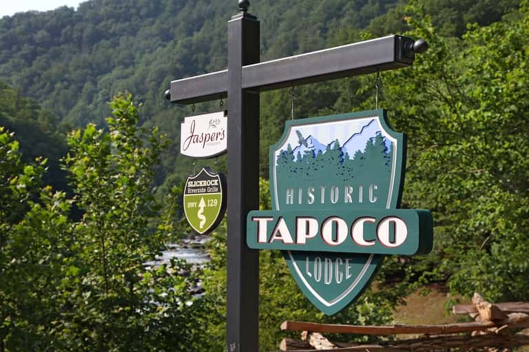Historic Tapoco Lodge sign with Jaspers restaurant sign hanging on black post in forested mountain valley setting