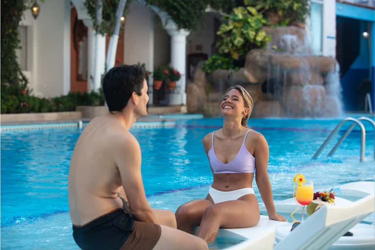 Couple enjoying pool at hotel with waterfall feature, tropical architecture, and blue umbrellas