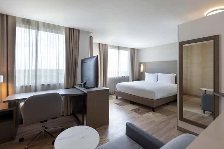Contemporary hotel suite with bed, work desk, TV, and floor-to-ceiling windows