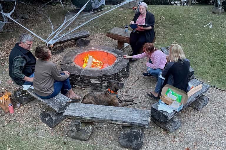 Family gathering around outdoor fire pit on grass with log seating and person reading nearby