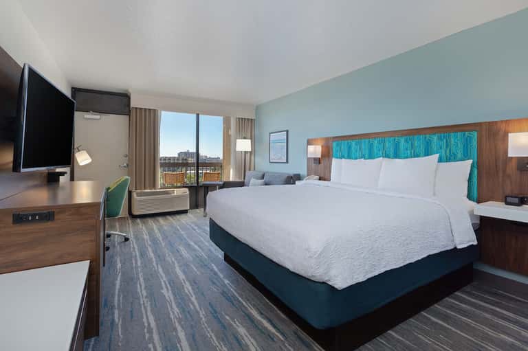 Hotel bedroom with turquoise accent headboard, white bedding, desk, TV, and city view windows
