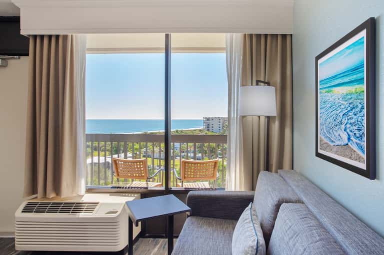 Hotel room with ocean view balcony, beige curtains, seating area, and framed beach artwork on light blue wall.