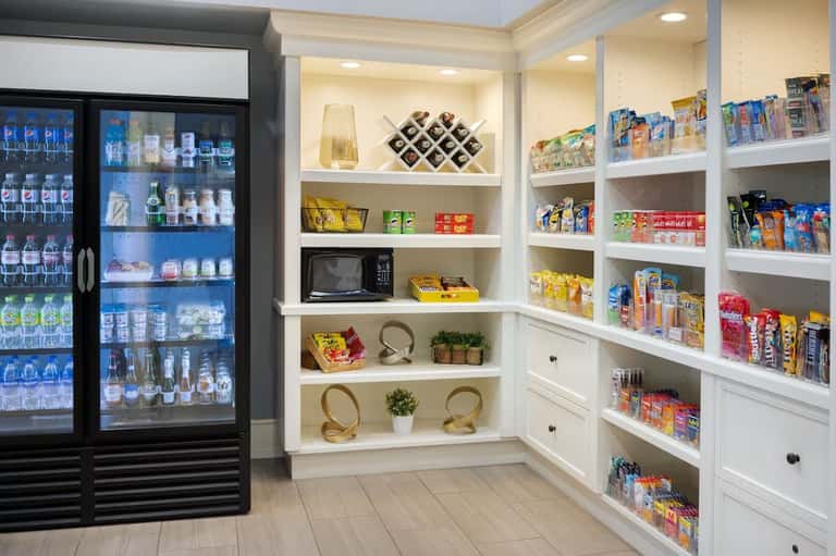 Hotel convenience store with refrigerated beverage cooler and white shelving stocked with snacks and products