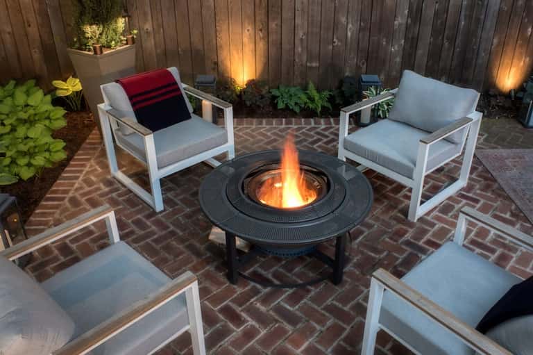 Brick patio with fire pit table, white modern chairs, gray cushions, and dark wooden fence at night