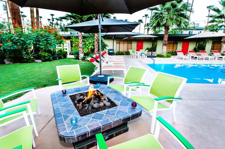 Resort pool area with lime green lounge chairs, blue umbrellas, tile fire pit, palm trees, and red-door cabanas