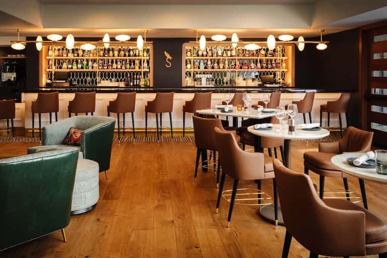 Upscale restaurant bar with wood flooring, brown leather seating, illuminated shelving, and elegant pendant lighting