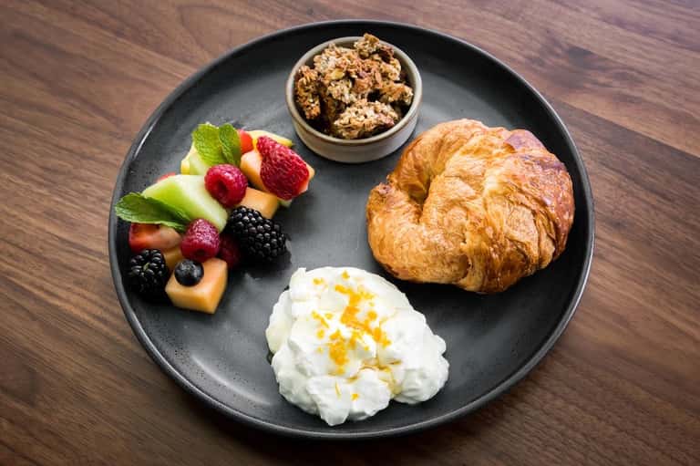Breakfast plate with croissant, fresh berries, granola, yogurt, and fruit on dark dish