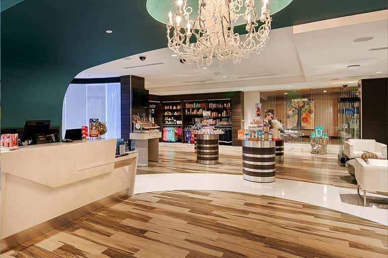 Modern hotel lobby with teal accent wall, crystal chandelier, wooden flooring, and retail shops
