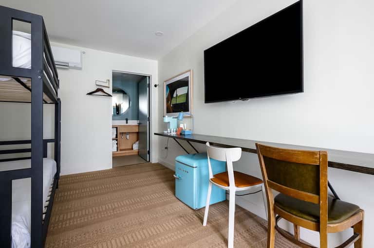Budget hotel room with bunk bed, desk with TV, bathroom access, and brown carpet flooring