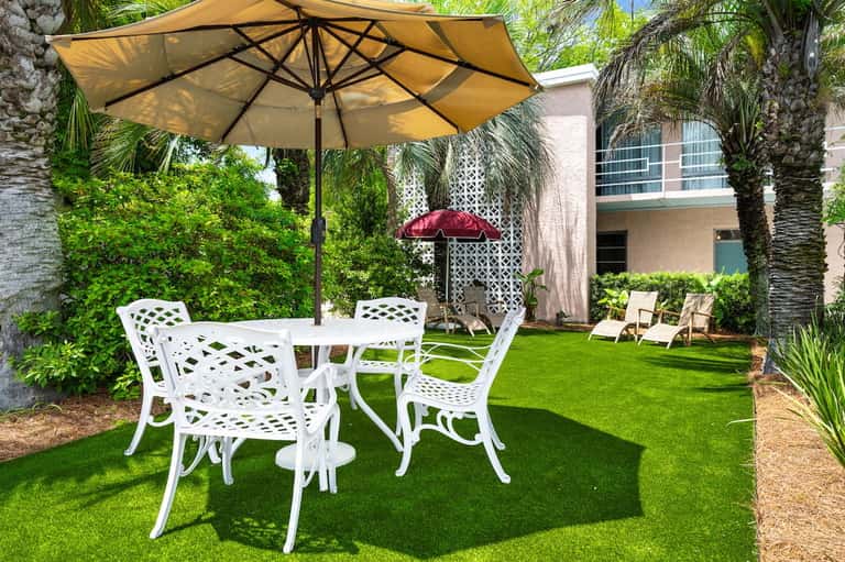 Backyard patio with white metal chairs, yellow umbrella, and manicured green lawn surrounded by palm trees