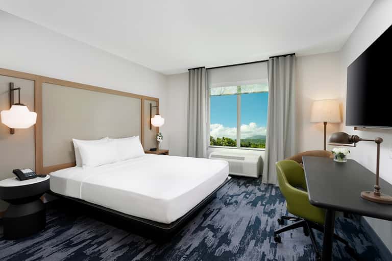 Modern hotel bedroom with queen bed, desk area, and large window overlooking mountains and blue sky